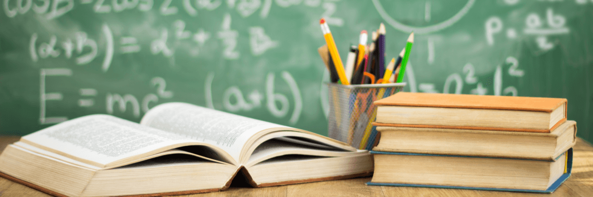 closeup of open textbook, cup of pens and pencils, and stack of books on a school or teacher desk, with a chalkboard with math and numbers behind, school or education industry concept