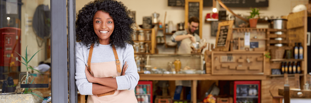 Portrait of a smiling young African American barista leaning with her arms crossed on the door of a trendy cafe, small business owner or SMB concept