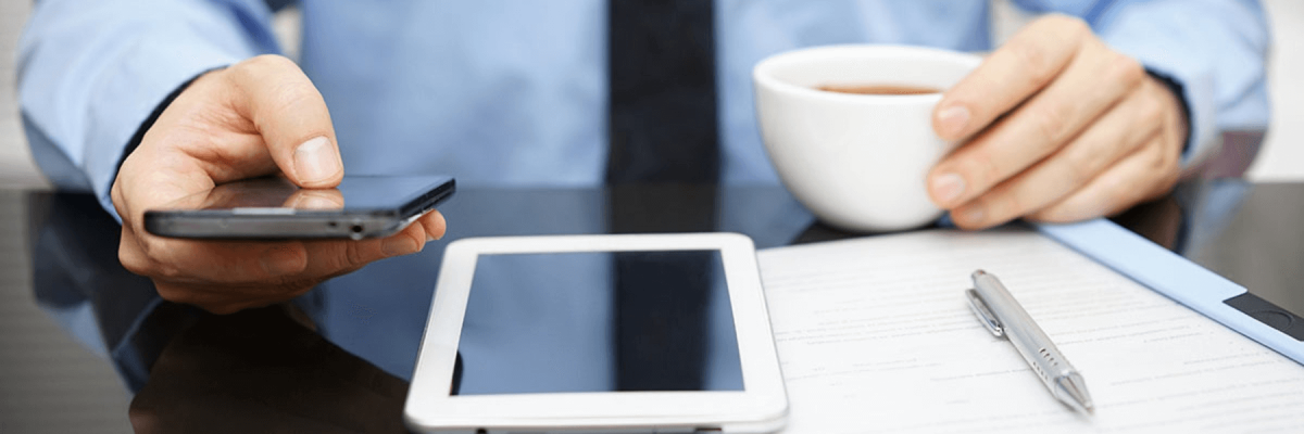 business man with cup of coffee in one hand, cell or mobile phone in the other, with a tablet and notebook or papers on the desk
