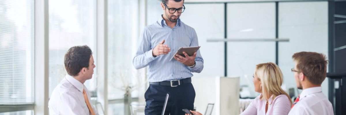 business team or partners standing around and sitting at a conference table, center focus on one man standing holding a clipboard or tablet